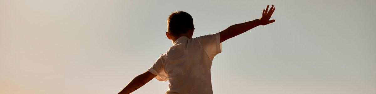 Silhouette of father carrying son on his shoulders at sunset. Rear view of unknown man and child pretending they can fly with their arms outstretched. Single parent bonding with his little boy outside